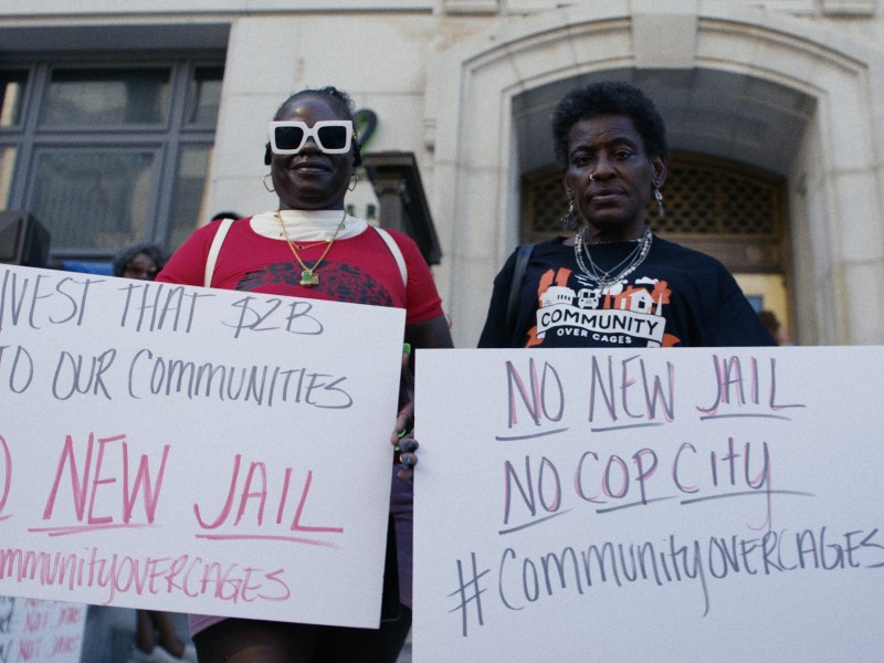 Two advocates hold signs outside protesting the Atlanta City Detention Center lease outside the Fulton County Courthouse. The sign on the left says, "Invest that $2B in our communities. NO NEW JAIL." The sign on the right says, "NO NEW JAIL. NO COP CITY"
