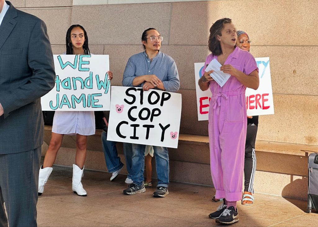 Image of a press conference just outside the Charlotte-Mecklenberg Courthouse during the morning hours. 

Jamie Marsicano, a white non-binary person with medium length dark blonde hair wearing a pink jumpsuit, stands in front of the supporters holding signs. Two of the signs are visible and read "We stand with Jamie," and "Stop Cop City."

One sign holder is a woman wearing rhinestone cowboy boots (she's going to a Beyonce concert later that evening and is dressed for such). The second visible signholder is an Asian American Pacific Islander man wearing blue jeans and a blue button down. A third supporter holds a sign that is obscured behind Jamie. 

Jamie and all three of her supporters are looking off to the right from the perspective of the camera. 

In the front left foreground, one of the attorneys representing Jamie that day stands in a blue suit, with his face cut off from the image. 
