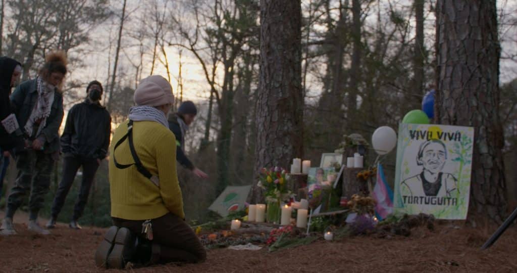 Photo by Lev Omelchenko A vigil attendee kneels next to a memorial built for Tortuguita.