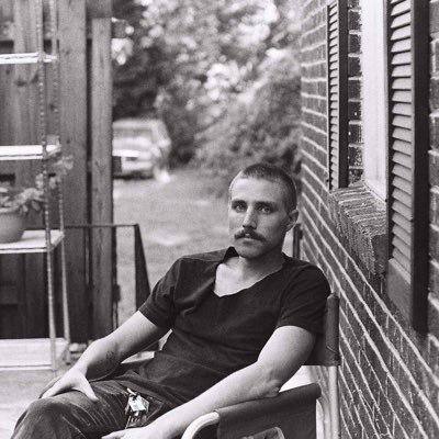 A black and white photo of Richard. He is sitting outside in a chair in front a brick wall. He is a slender white masculine person with closely cropped hair and a moustache.