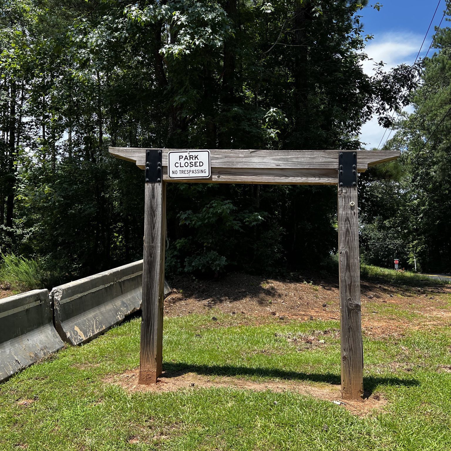 The remains of a sign that previously welcomed visitors to Intrenchment Creek Park. The empty wooden frame of the sign stands near where the new barricades were placed. A generic sign reading "PARK CLOSED  NO TRESPASSING" has been nailed to the top of the wooden frame.