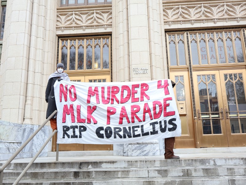Image of activists holding a banner that reads "No Murder 4 MLK Parade RIP Cornelius" in front of the City Hall Tower in Atlanta during daylight hours. The photo is shot from a lower angle, up concrete stairs. The activists stand in front of a marble and concrete facade with bronze doors and large windows. "City Hall" is written above the doors. An activist holding up the left side of the banner stands on a handrail. They hold the banner with a gloved hand and a wearing a peacoat, keffiyah, medical mask and beanie. The second activist is hidden behind the banner, but brown pants and shoes as visible below the banner and the front end of a hooded jacket is visible on the right side of the banner.