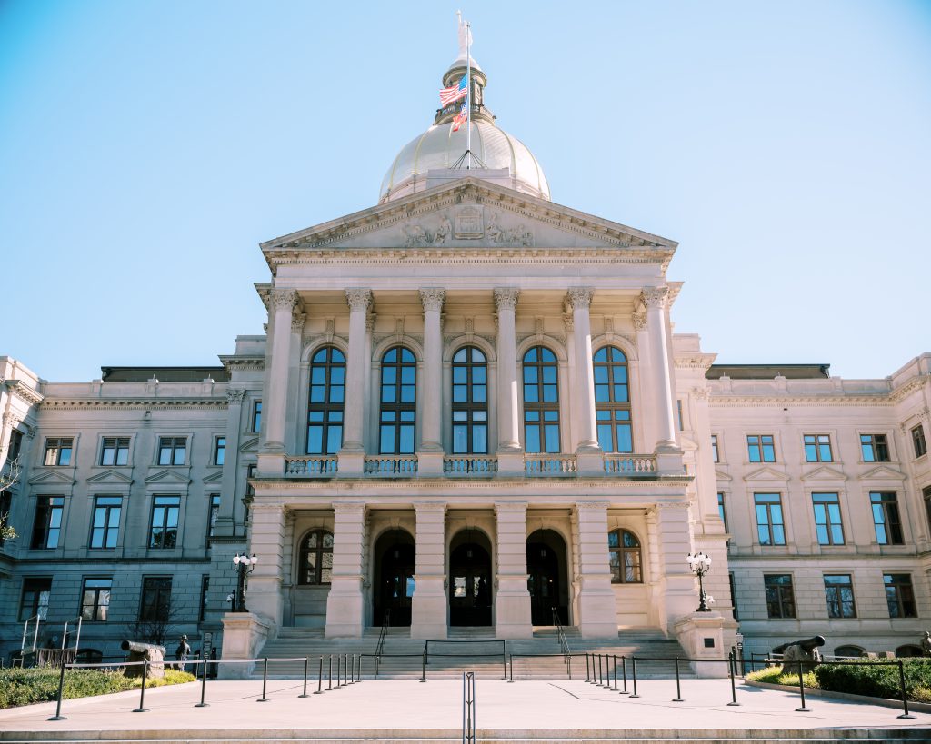 Image of the front facade of the Georgia State Capitol, known as the Gold Dome. The camera is facing the facade from a lower angle with the capitol steps in the bottom of the frame. Behind the gold dome, a blue sky with very light clouds is visible.