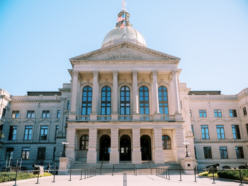 Image of the front facade of the Georgia State Capitol, known as the Gold Dome. The camera is facing the facade from a lower angle with the capitol steps in the bottom of the frame. Behind the gold dome, a blue sky with very light clouds is visible.