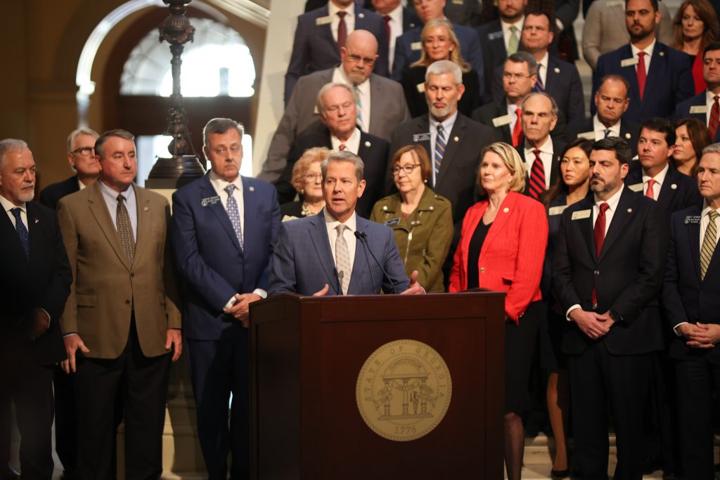 A group of Georgia legislators in business attire stand behind Governor Brian Kemp, who in turn stands behind a dark-wood podium with the state seal of georgia on the front. The group is situated in front of and on the stairs in the rotunda of the Georgia Capitol Building.