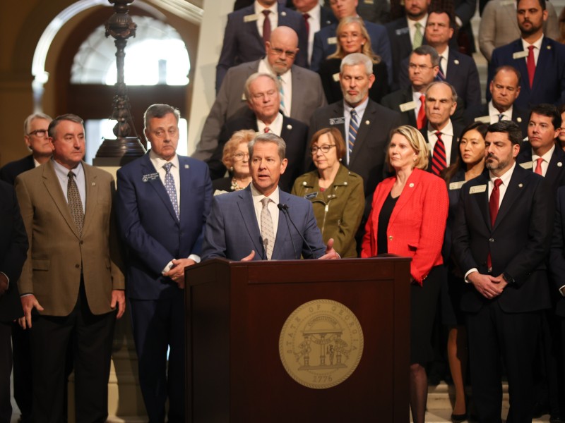 A group of Georgia legislators in business attire stand behind Governor Brian Kemp, who in turn stands behind a dark-wood podium with the state seal of georgia on the front. The group is situated in front of and on the stairs in the rotunda of the Georgia Capitol Building.