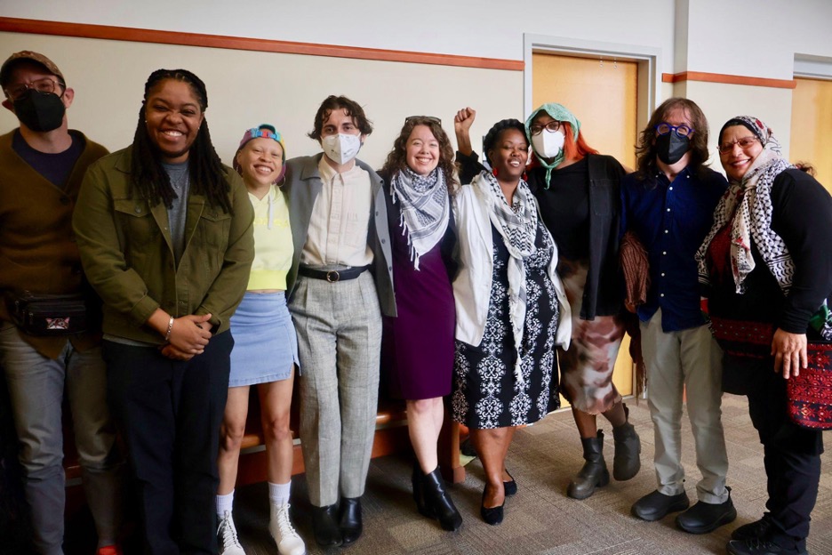 A group of nine smiling people wearing business casual attire stands in a curved line inside a beige hallway. The group is of mixed genders and races. The three individuals in the middle are the "Weealune Three" Cop City protesters. Left to right they are Noah Grigni wearing grey slacks, a pink oxford, grey jacket and KN-95 mask; Krya Hanon, wearing a purple dress and a keffiyeh, and Rukia Rogers, wearing a patterned black dress and keffiyeh. 