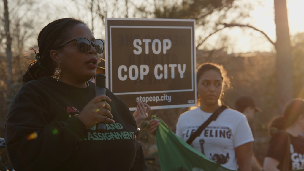Foreground: Atlanta Organizer Mary Hooks speaks over a microphone as a fellow organizer holds a sign that says "Stop Cop City" in the background.