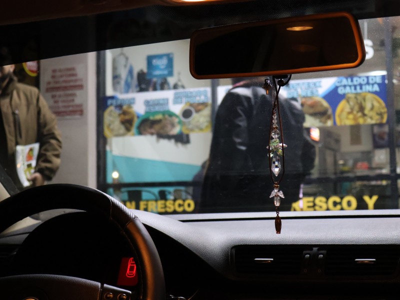 Photo of the inside of a car's windshield in the dark. In the foreground, a crystal angel hangs from the car's rearview mirror. In the background, ICE Chasers holding Know Your Rights materials stand in front of a restaurant with breakfast items in Spanish.