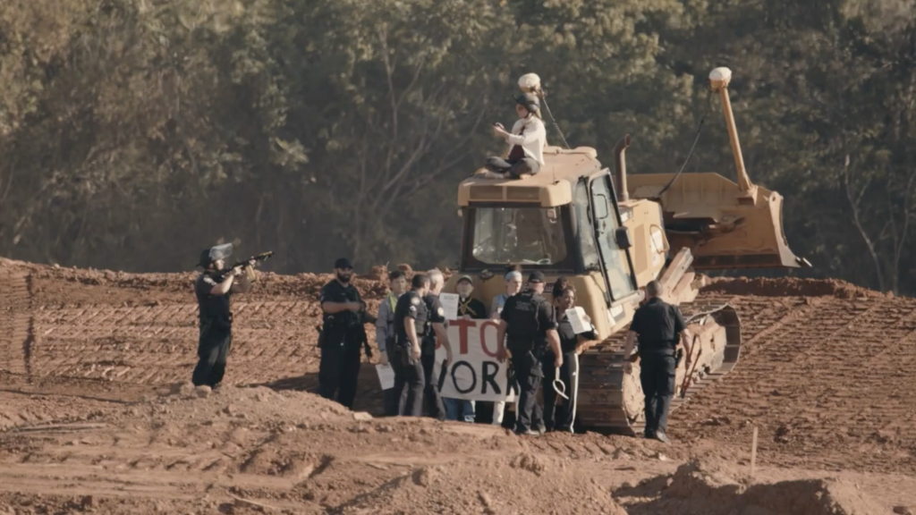 Activists, including faith leaders, chained to a bulldozer. One activist sits on top. Police move in to arrest the activists. One officer is pointing a weapon at them.