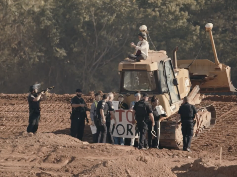 Activists, including faith leaders, chained to a bulldozer. One activist sits on top. Police move in to arrest the activists. One officer is pointing a weapon at them.