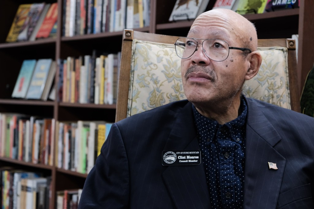 Clint Monroe, an older Black man with a bald head, glasses and a mustache, looks off to the left frame of the camera. Monroe wears a dark, feintly pinstripe suit and a blue dotted button down shirt. He has a City of Stone Mountain Council Member name tag on his right lapel and a small american flag pin on his left lapel. 

Monroe sits on a wooden chair with a beige floral pattern behind him. Bookshelves sit in the background. 