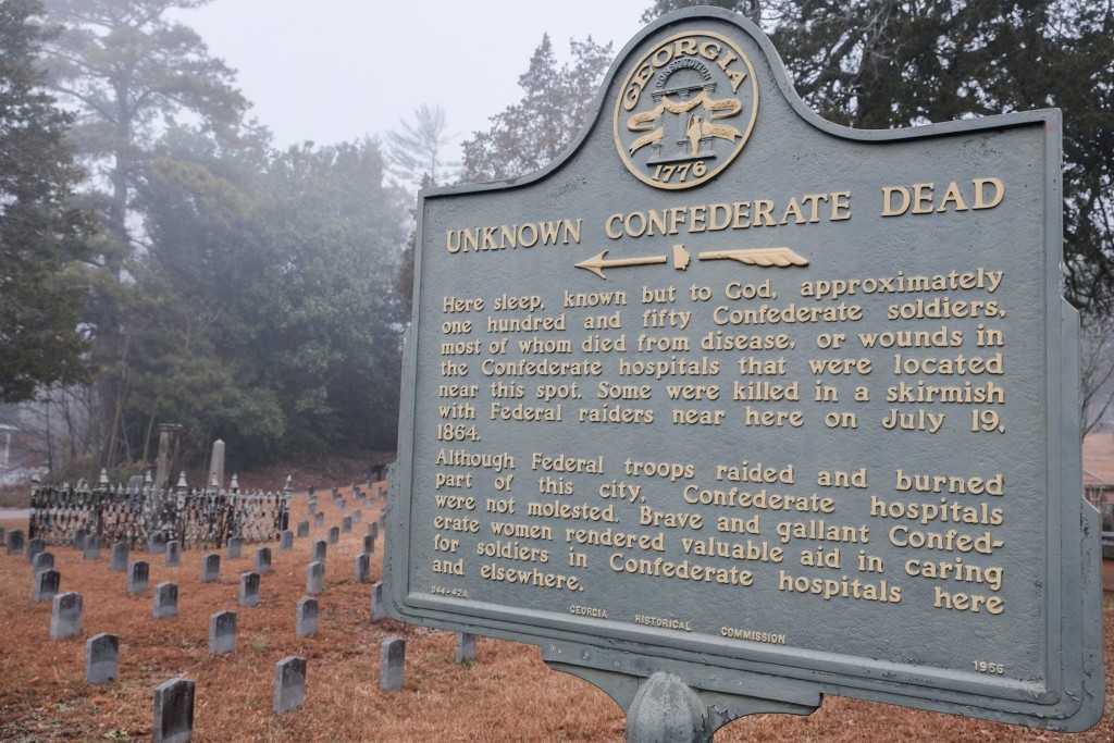 A bronze historical marker—turned green by age—with gold-colored lettering sits in the foreground and in the right of the frame. The marker has the state seal of Georgia at the top. The title reads, "Unknown Confederate Dead." The caption reads "here sleep, known but to God, approximately one hundred and fifty Confederate soldiers, most of whom died from disease, or wounds in the Confederate hospitals that were located near this spot. Some were killed in a skirmish with Federal raiders near here on July 19, 1864. Although Federal troops raided and burned part of this city, Confederate hospitals were not molested. Brave and gallant Confederate women rendered valuable aid in caring for soldiers in Confederate hospitals here and elsewhere." In small text at the bottom reads "Georgia Historial Commission 1956" It is a cloudy day. Behind the historical marker sit granite gravestones, weathered with age. Each is marked "CSA"