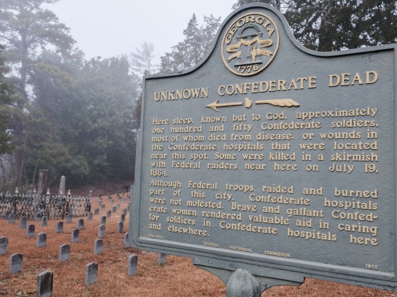 A bronze historical marker—turned green by age—with gold-colored lettering sits in the foreground and in the right of the frame. The marker has the state seal of Georgia at the top. The title reads, "Unknown Confederate Dead." The caption reads "here sleep, known but to God, approximately one hundred and fifty Confederate soldiers, most of whom died from disease, or wounds in the Confederate hospitals that were located near this spot. Some were killed in a skirmish with Federal raiders near here on July 19, 1864. Although Federal troops raided and burned part of this city, Confederate hospitals were not molested. Brave and gallant Confederate women rendered valuable aid in caring for soldiers in Confederate hospitals here and elsewhere." In small text at the bottom reads "Georgia Historial Commission 1956" It is a cloudy day. Behind the historical marker sit granite gravestones, weathered with age. Each is marked "CSA"