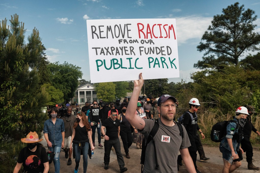 A group of protesters walk along a dirth path with green foliage and trees on either side. In the background is an Antebellum Era building with plantation architecture. The protesters are wearing mostly dark clothing, one is in a Black Lives Matter t-shirt. In the foreground and center of the picture is a white male protesters wearing on a grey shirt a "Remove the Hate" sticker that has a red Anarchy A on it. He holds a white poster board at a landscape angle that reads "Remove racism from our taxpayer funded public park."
