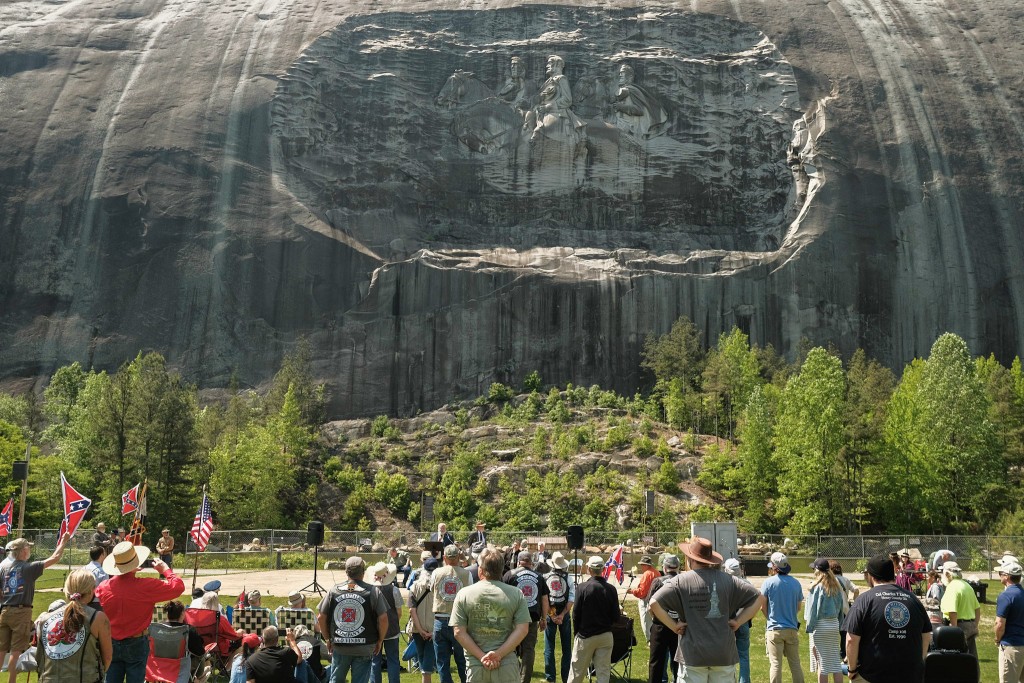 A group of white people stands and sit at the bottom of the frame. Some hold Confederate flags or wear confederate supportive clothing. In the background, dominating the upper half of the photo is the portion of Stone Mountain—a granite mountain— with a relief carved into it. The relief features Confederate Leaders Jefferson Davis, Gen. Robert E. Lee, and Gen. Thomas J. "Stonewall" Jackson riding on horseback.