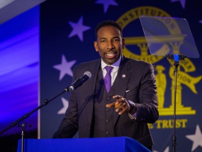 Andre Dickens stands on a stage in dark lighting with a hint of blue. He is wearing a dark suit and vest with a purple tie and light colored shirt. He is pointing and looking at the camera. He stands behind a blue podium. The upper left corner of the Georgia State Flag is visible behind him.