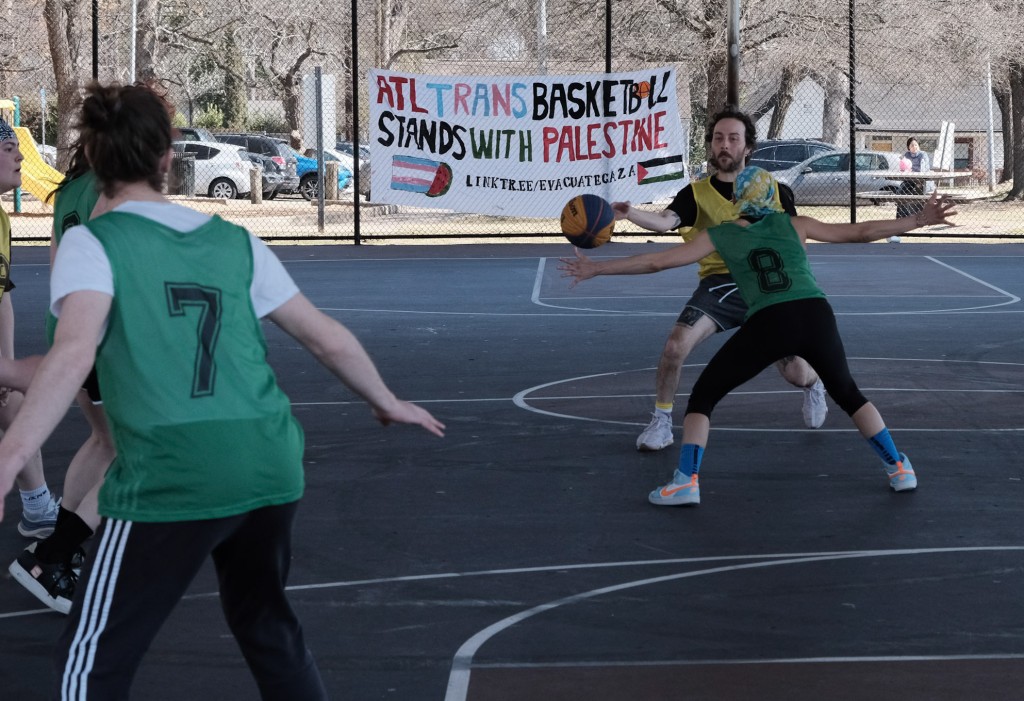 A basketball player wearing yellow dribbles the ball while another player in green blocks them. A sign reading, "ATL Trans Basketball Stands With Palestine" is hung in the background.