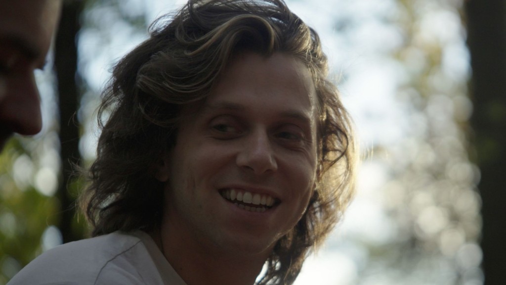 A cinematic still of Jamie Mariscano, a white nonbinary person with medium-length dark blonde hair wearing a white t-shirt. Jamie is smiling. The setting is close to dusk, with long shadows. The sun frames left side of Jamie's face.

Only the upper quarter of Jamie is visible.  

The setting is woodsy, with trees visible but slightly blurry in the background. The 