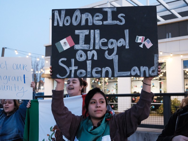 A woman stands at a rally holding a sign that says, "No one is illegal on stolen land."