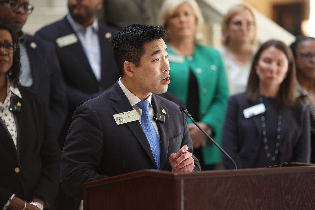 Georgia State Rep. Sam Park stands behind a podium during a press conference in the Georgia State Capitol Building. A group of Democratic legislators stand behind him. 
