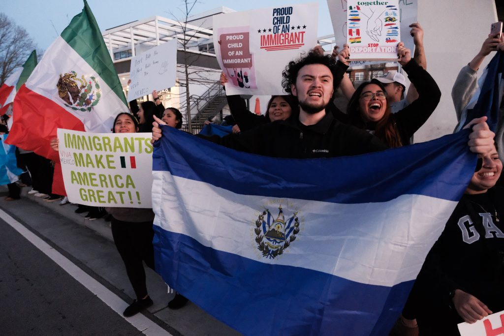 A group of smiling protestors hold up signs in support of immigration, reading "Immigrants Make America Great" and "Proud Child of an Immigrant." The subject of the photo, a man standing at the front of the crowd, holds up the flag of El Salvador.