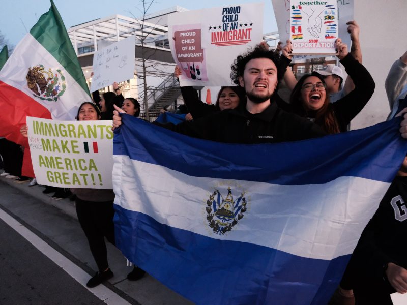 A group of smiling protestors hold up signs in support of immigration, reading "Immigrants Make America Great" and "Proud Child of an Immigrant." The subject of the photo, a man standing at the front of the crowd, holds up the flag of El Salvador.