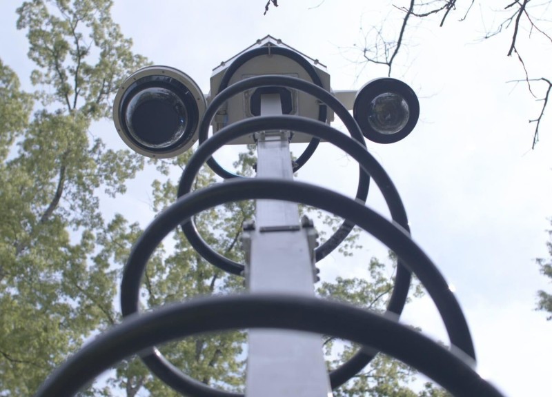 A device with two cameras, part of the Atlanta surveillance camera network, stands on a pole against a cloudy sky backdrop with trees in the background.