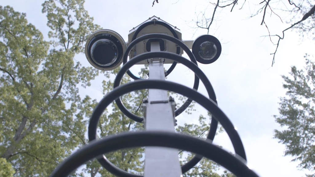 A device with two cameras, part of the Atlanta surveillance camera network, stands on a pole against a cloudy sky backdrop with trees in the background.