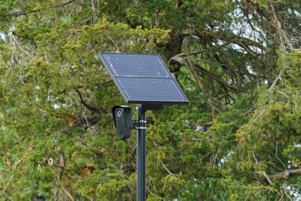 A Flock Safety Camera, which have been used to track license plates on behalf of ICE, sits center frame with a backdrop of trees.