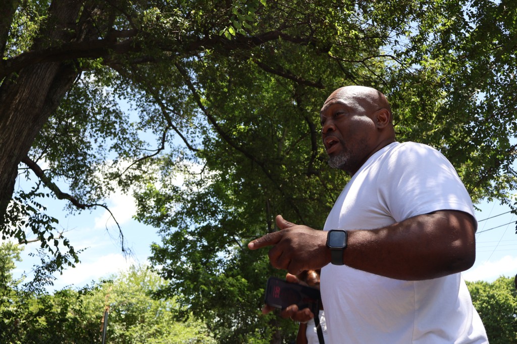 A man standing and speaking on the righthand side of the frame with trees in the background.