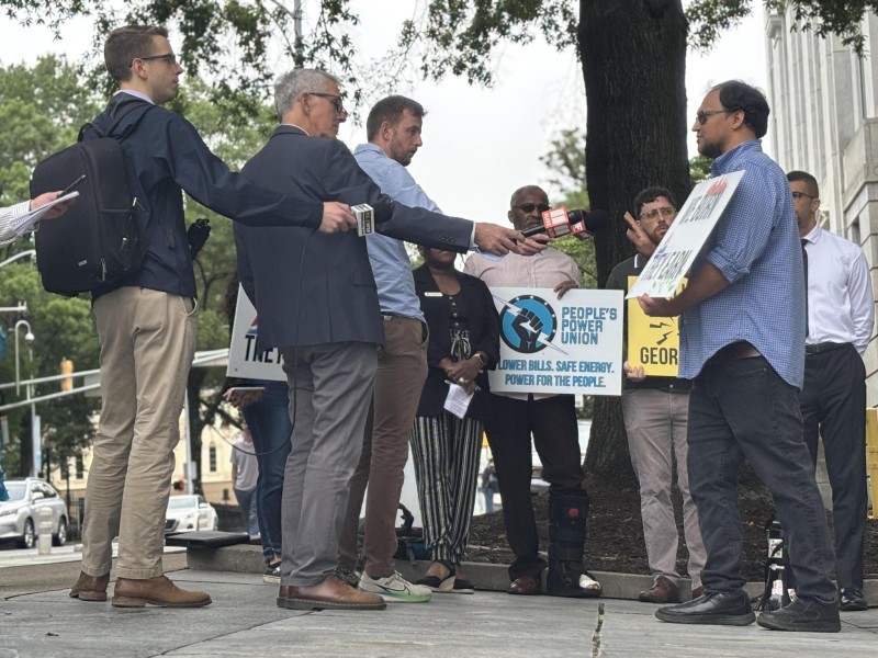 Two reporters in profile speaking with a man holding a sign that has Georgia Power's logo on it and reads "We Burn They Earn."