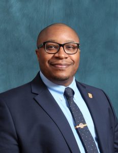 A headshot of Marshall Freeman, wearing a navy blue suit, light blue oxford, and blue patterned tie. 