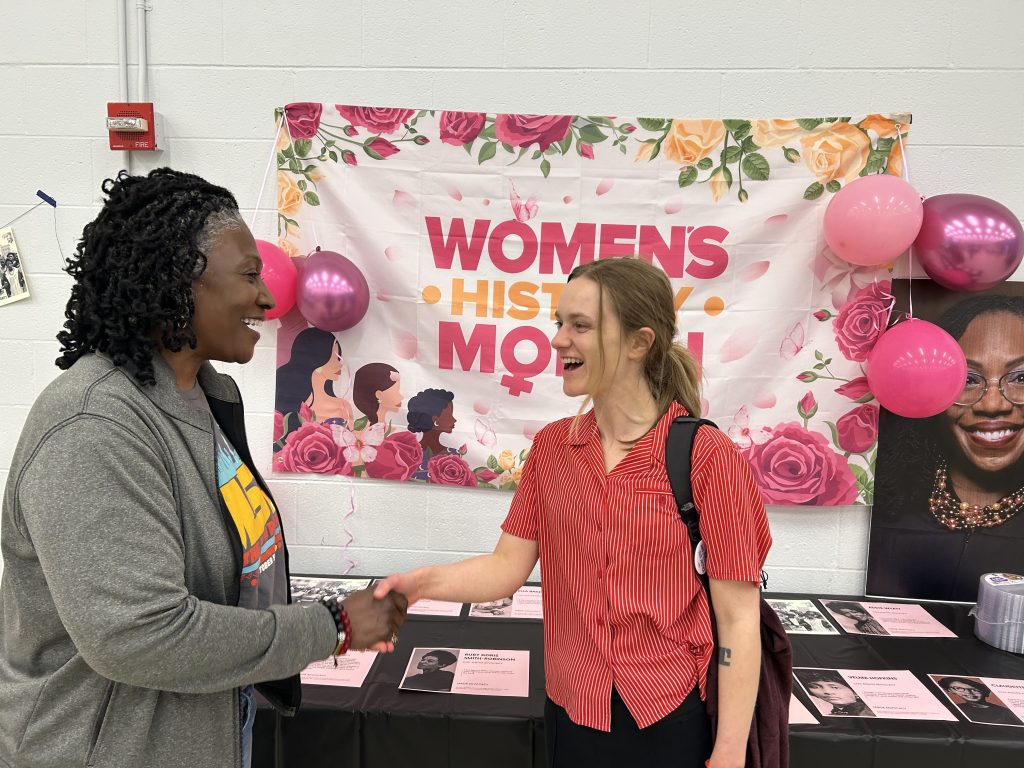A woman and a nonbinary femme-presenting person shaking hands in front of of a pink "Women's History Month" poster.