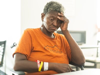 An older Black woman in an orange shirt sits at a table and holds her head in her left hand.