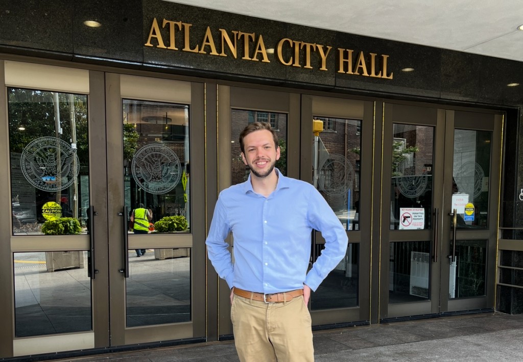 A man in a powder blue button-down and khakis stands in front of a pair of doors. Above the doors, there are letters that read "Atlanta City Hall."