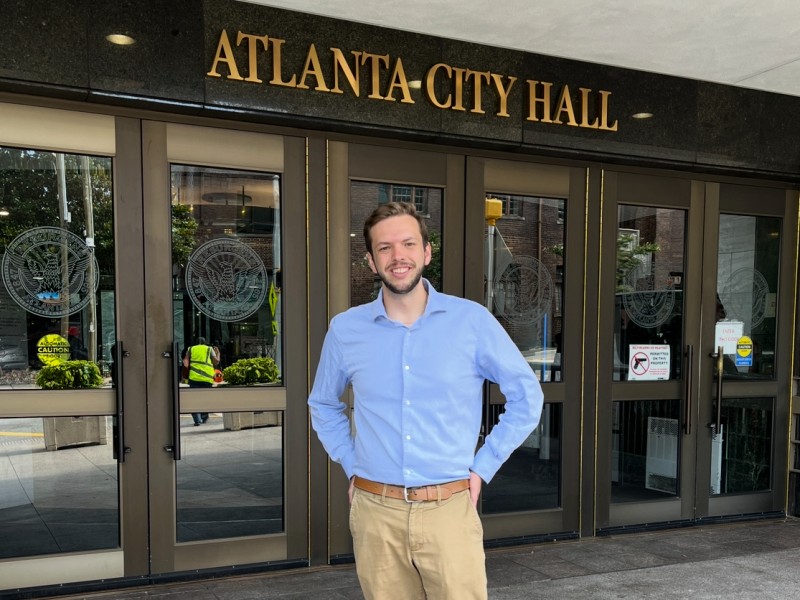A man in a powder blue button-down and khakis stands in front of a pair of doors. Above the doors, there are letters that read "Atlanta City Hall."