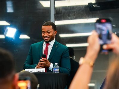 A man in a suit standing at a lectern with a name card that reads "Andre Burgin." Two people in front of him are recording a video of him on their phones.