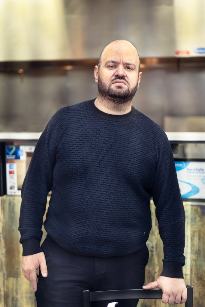 A bald man with a beard standing in an arepa restaurant and wearing all black.