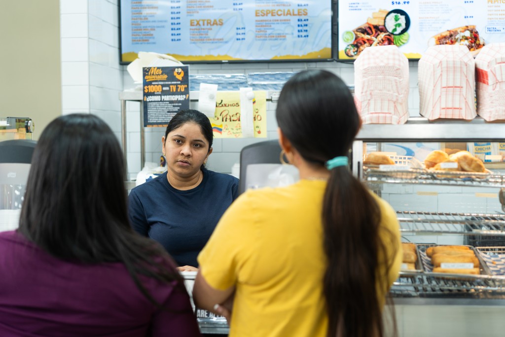 Two women with their backs to the camera order off a menu on a TV above their heads. Another woman—the cashier—looks at them as they make their decisions.