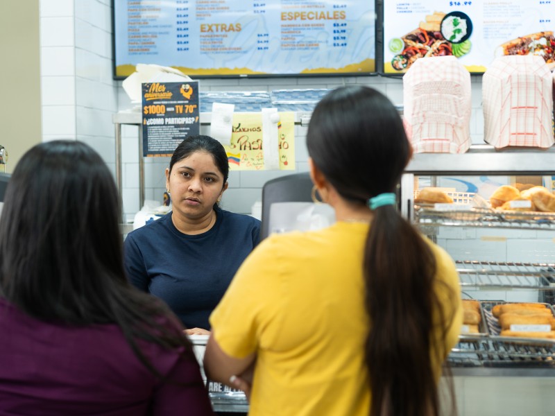 Two women with their backs to the camera order off a menu on a TV above their heads. Another woman—the cashier—looks at them as they make their decisions.