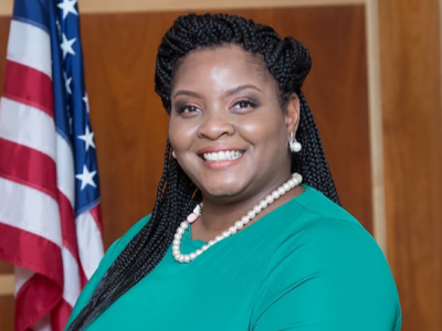 A woman with braids and wearing pearls smiles into the camera. An American flag is behind her.