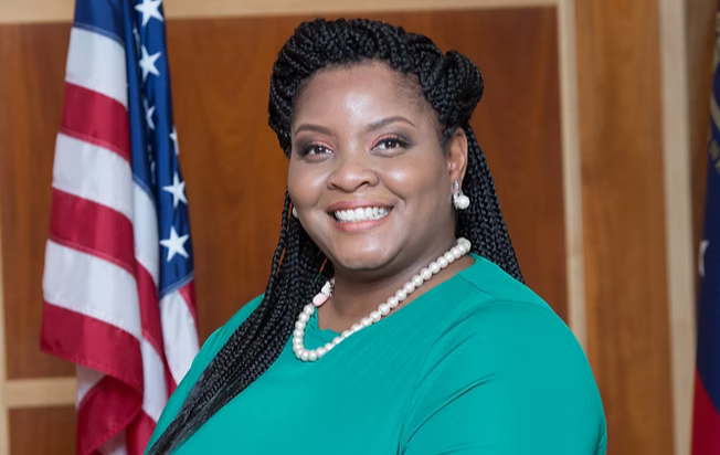 A woman with braids and wearing pearls smiles into the camera. An American flag is behind her.