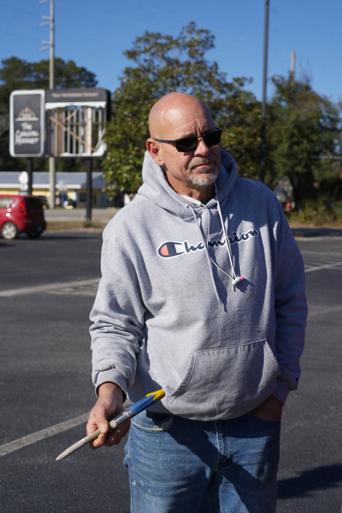 A bald man with sunglasses and a goatee holds a paintbrush.