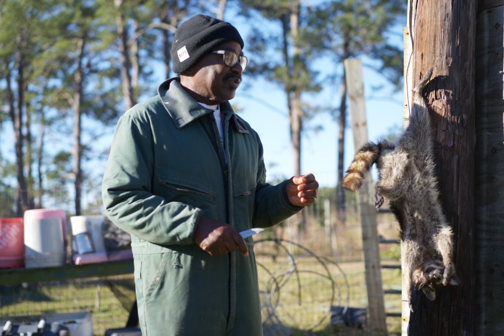 A man wearing glasses and a beanie stands next to a raccoon carcass.