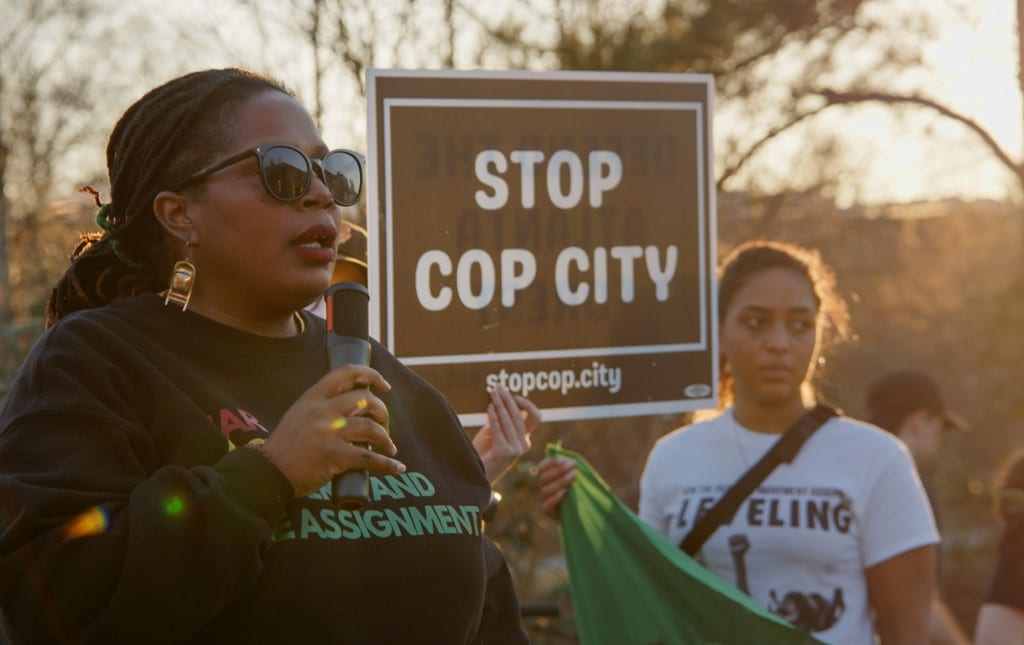 Foreground: Atlanta Organizer Mary Hooks speaks over a microphone as a fellow organizer holds a sign that says "Stop Cop City" in the background.