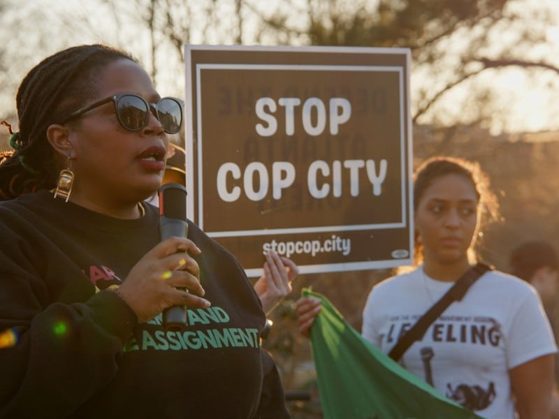 Foreground: Atlanta Organizer Mary Hooks speaks over a microphone as a fellow organizer holds a sign that says "Stop Cop City" in the background.